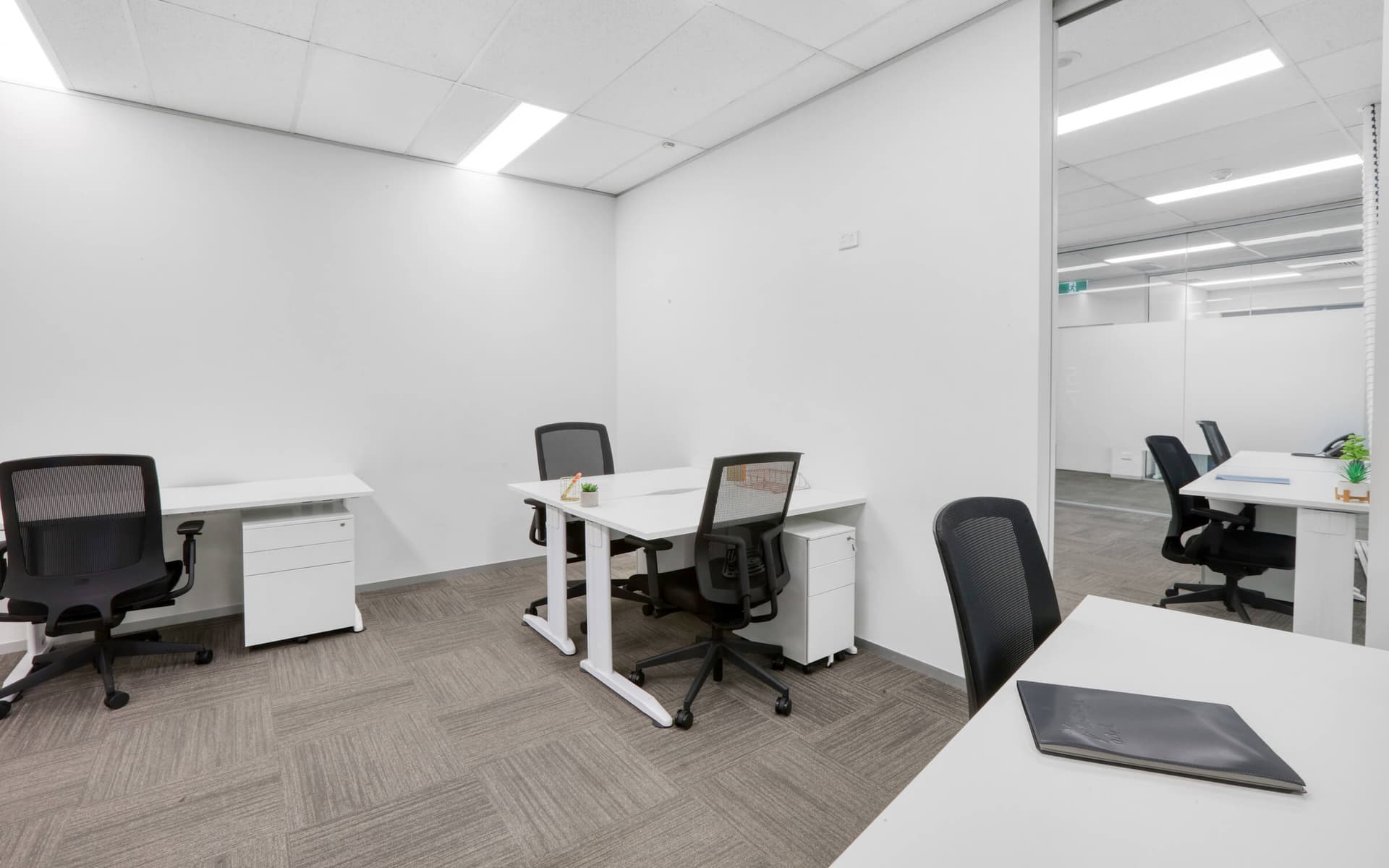 Modern office space with white desks, black mesh chairs, and neutral carpet. Clean, minimalist design.