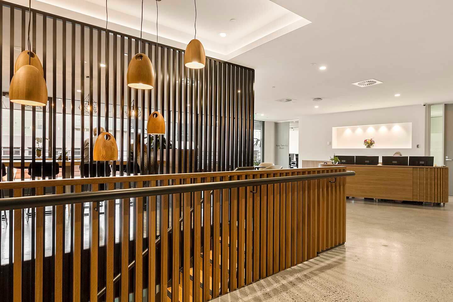 Modern office interior with a wooden railing and hanging pendant lights. A reception desk is visible in the background with a floral arrangement. The space features a partition with vertical wood slats and polished concrete floors.