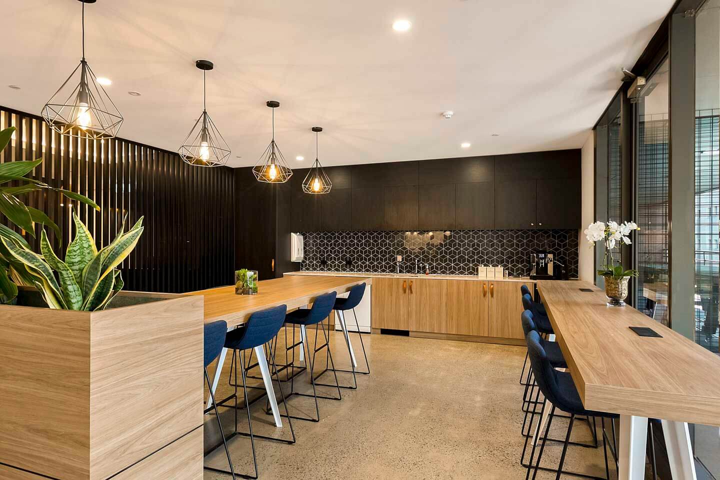 Modern kitchen with wooden cabinetry and black accents. Features long island with blue chairs, pendant lighting, and geometric backsplash. Large plants decorate the space, and natural light pours in through floor-to-ceiling windows on the right.