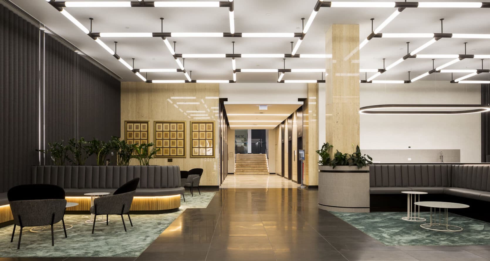 Modern lobby with a sleek design, featuring dark tiled floors and cushioned seating areas alongside small round tables. Vertical lighting fixtures grace the ceiling, while plants add a touch of nature in corners. Perfect for shared office space in Sydney, with a stylish staircase in the background.