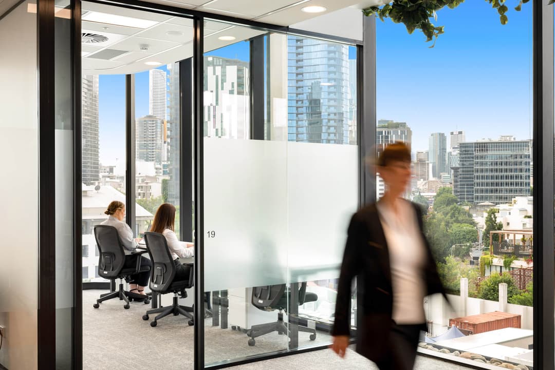 A modern office space with glass walls. Two people are seated at desks working on computers, while a blurred person walks past. Large windows reveal a city skyline with tall buildings and greenery outside.