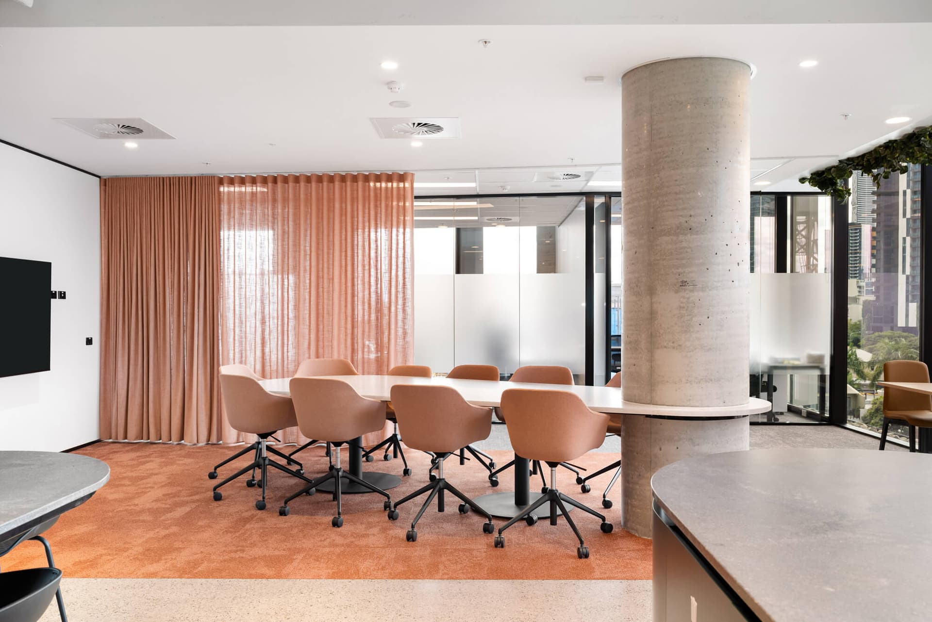 Modern office conference room with pink chairs, large table, and a textured concrete pillar. Glass walls and light-filled interior.