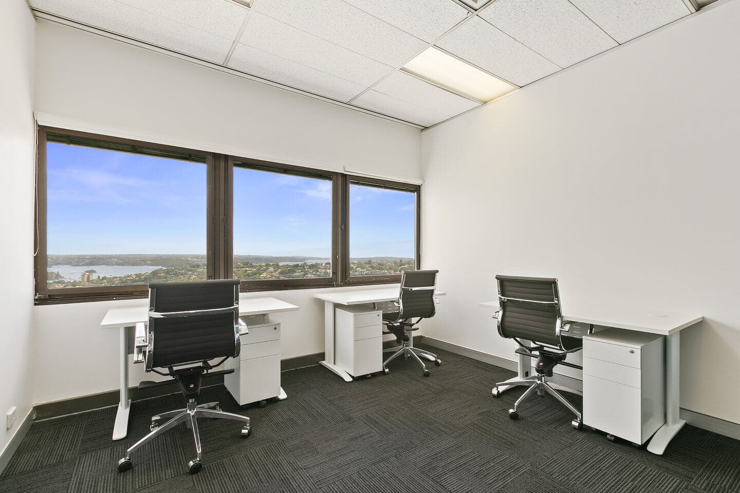 Office space with three black chairs and desks arranged against a large window with a view of a cityscape and water. The room features white walls, a dark carpet, and overhead fluorescent lighting.