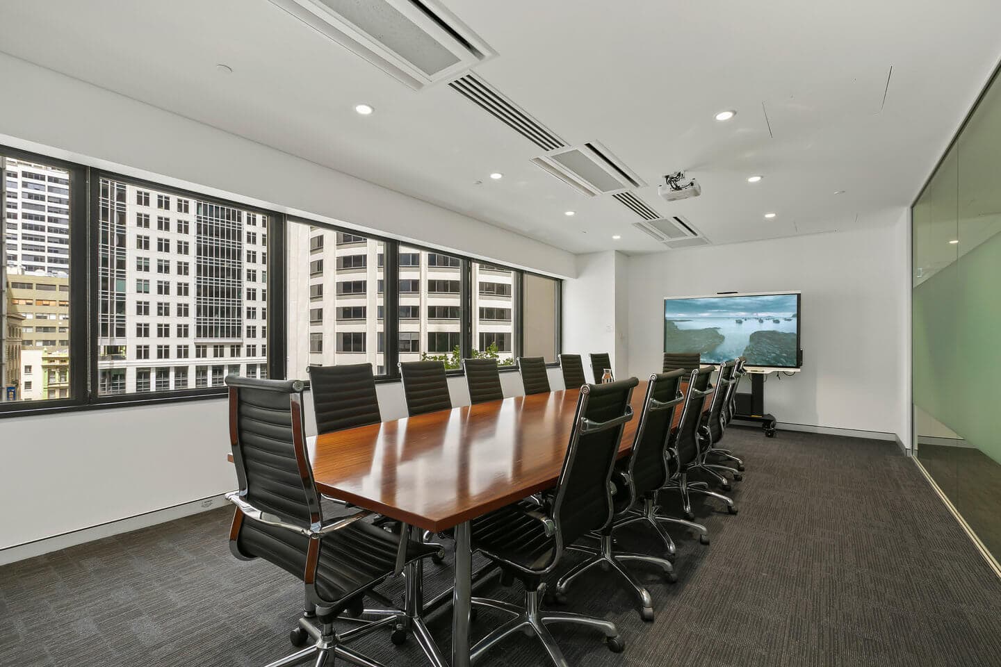 A modern conference room with a long wooden table surrounded by black leather chairs. Large windows offer a view of city buildings. A flat-screen TV is mounted on the wall at the far end displaying an ocean scene.