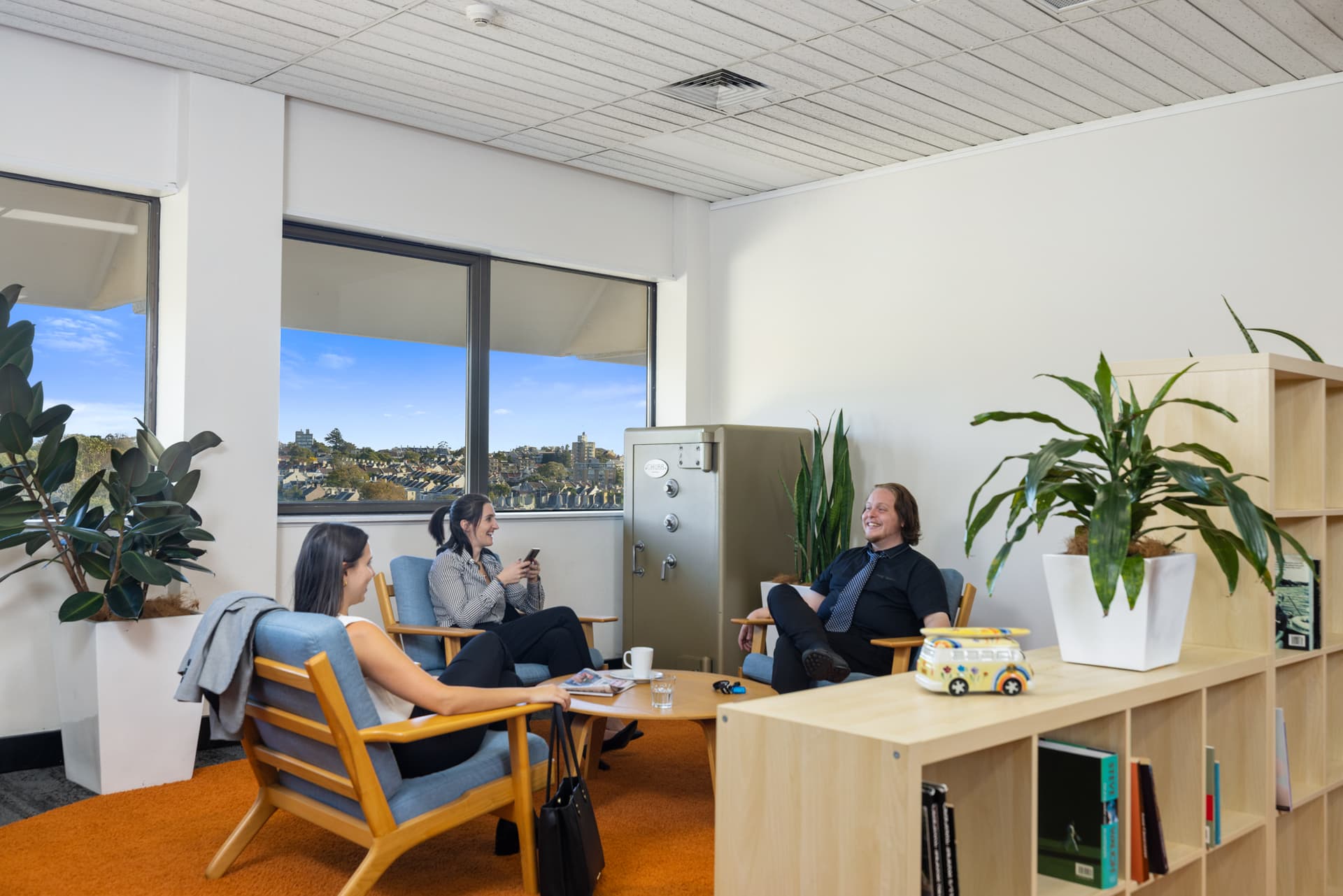 Three people sit and talk in a modern office lounge with large windows, indoor plants, a safe, and bookshelves. They appear relaxed, enjoying a casual conversation. Natural light fills the room.