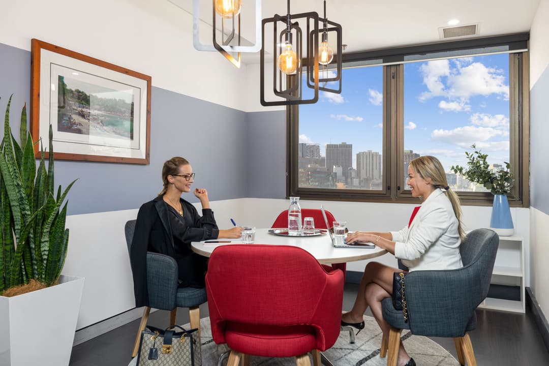 Two women sit across from each other at a round table in a modern office with large windows, red and gray chairs, and a plant in the corner. City buildings and blue sky are visible outside.