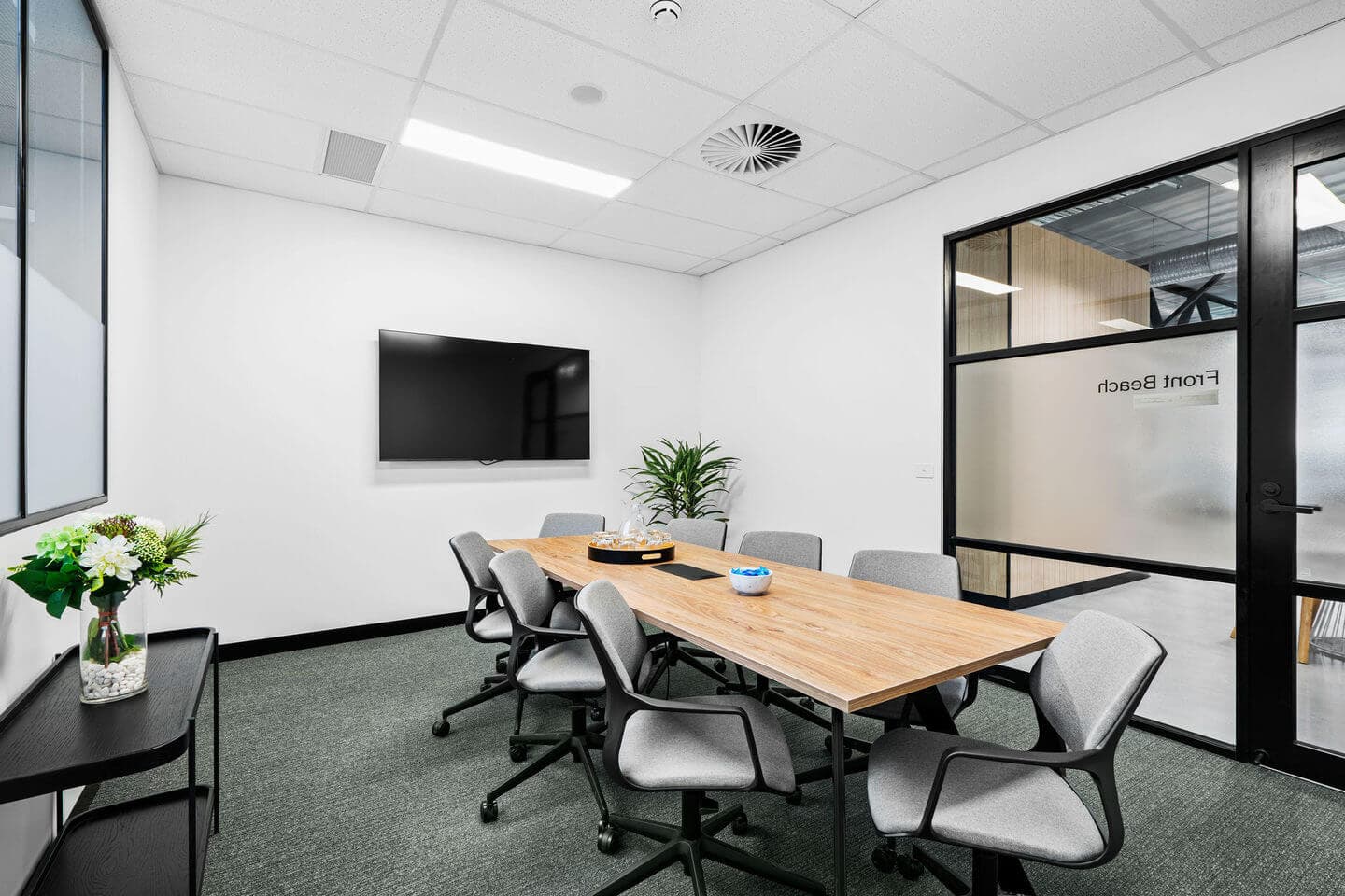 A modern conference room with a wooden table surrounded by six gray chairs. A flat-screen TV is mounted on the white wall. A plant and a vase with flowers decorate the space. Glass doors with a frosted section lead to an adjacent area.