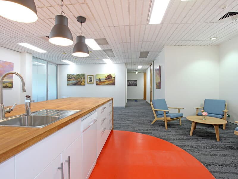 Modern office kitchen with a wooden countertop and stainless steel sink. Hanging pendant lights illuminate the area. The room has blue armchairs, a small table, and colorful art on the walls. Carpeted flooring with a vibrant orange section.