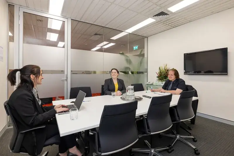 Three people are seated around a meeting table in a modern conference room. They are engaged in discussion. There is a laptop, papers, and a glass of water on the table. A flat-screen TV and a plant are visible on the wall.