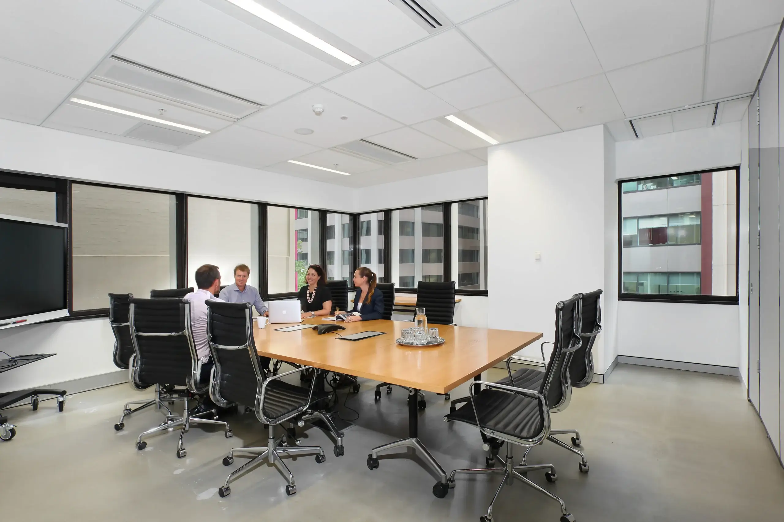 Four people are seated around a large wooden conference table in a modern office setting. The room has white walls, black chairs, and large windows providing natural light. A TV screen is mounted on the wall.