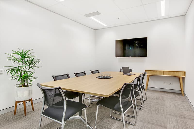 Small, modern conference room with a rectangular wooden table surrounded by black chairs. A flat-screen TV is mounted on the white wall. A potted plant is in the corner, and a small wooden console table sits against the wall.