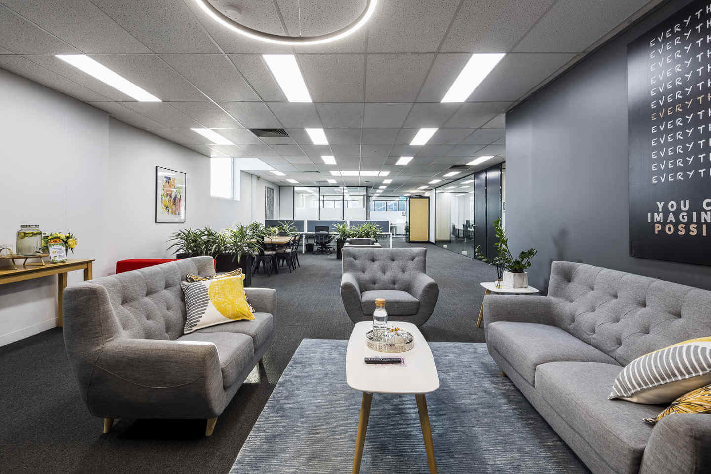 Modern office lounge with gray sofas, a white coffee table, and a rug. Decor includes plants, a wall poster, and a motivational sign. A dining table is visible in the background under bright lighting.