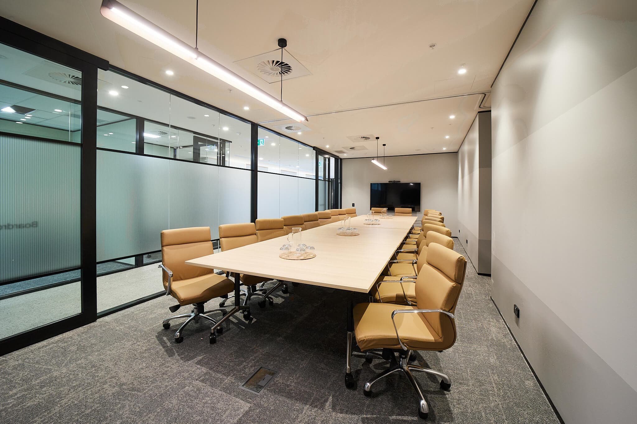 A modern conference room with a long wooden table surrounded by tan leather swivel chairs. The room features frosted glass walls, ceiling lights, a television screen on one end, and a neutral color palette.