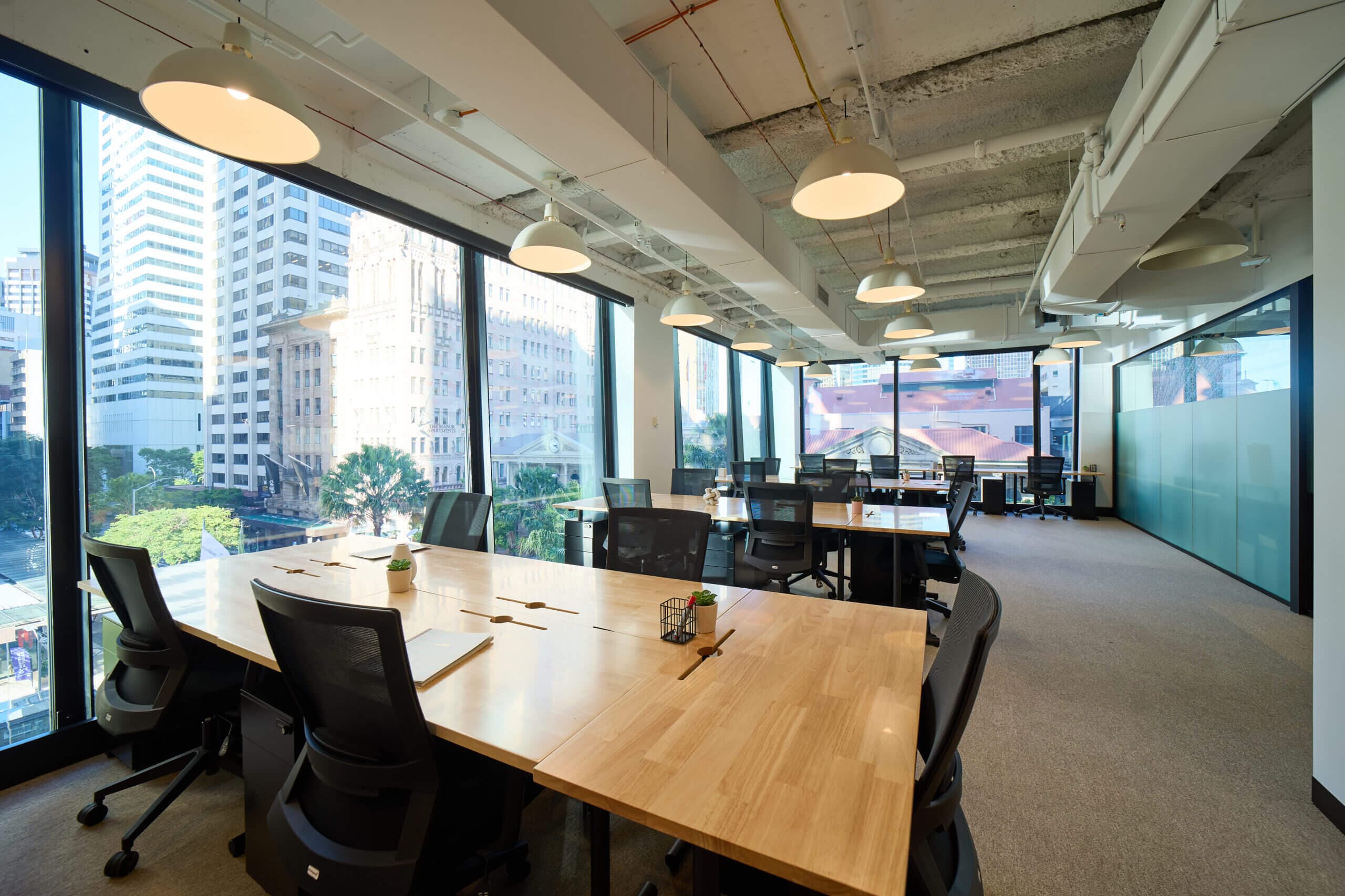 A modern office space with large windows offering a city view. There are wooden desks with black office chairs, ceiling lights, and a meeting room in the background. The room is well-lit and has a professional atmosphere.