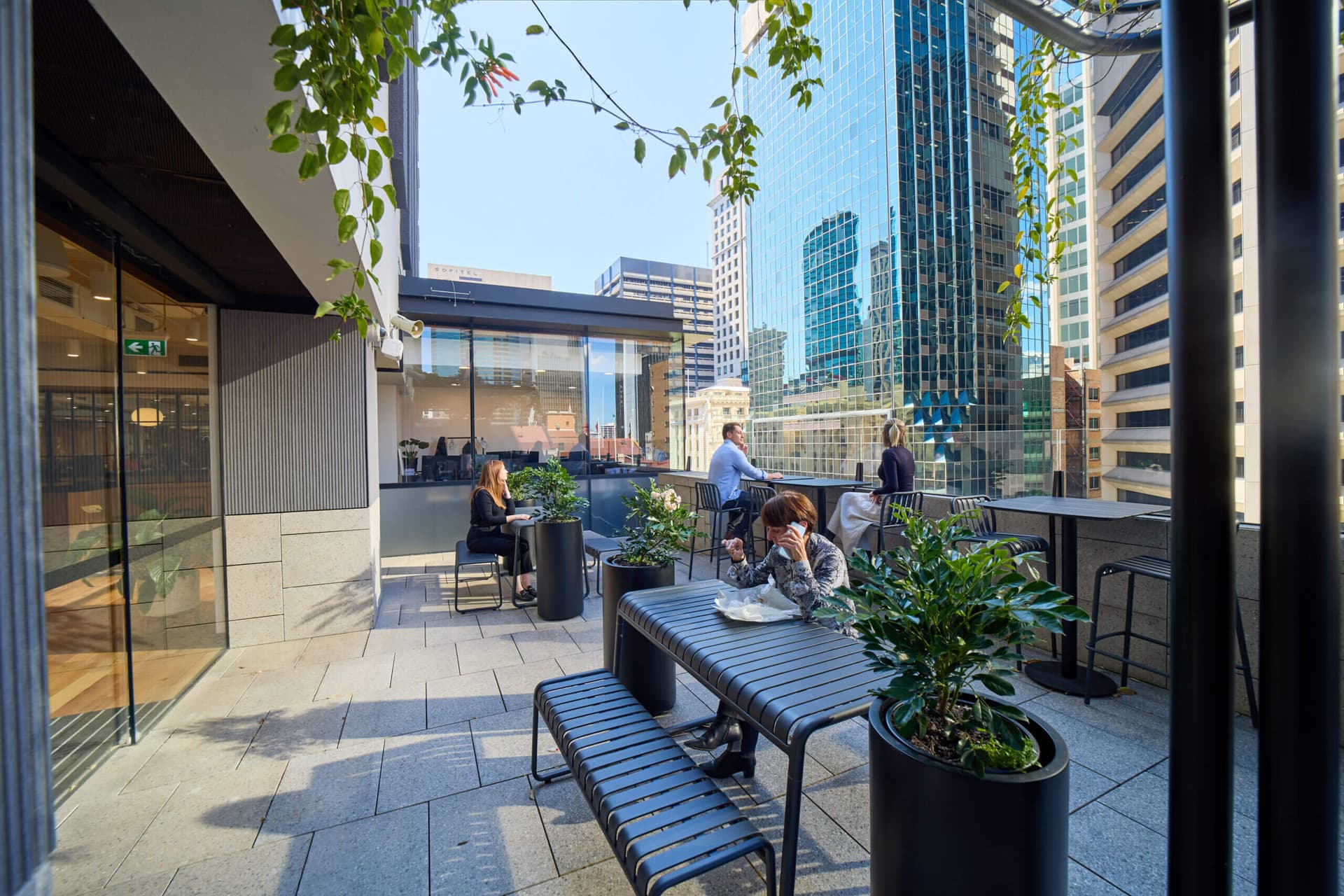 Outdoor terrace seating area with people eating, working, and talking. City buildings visible in background.