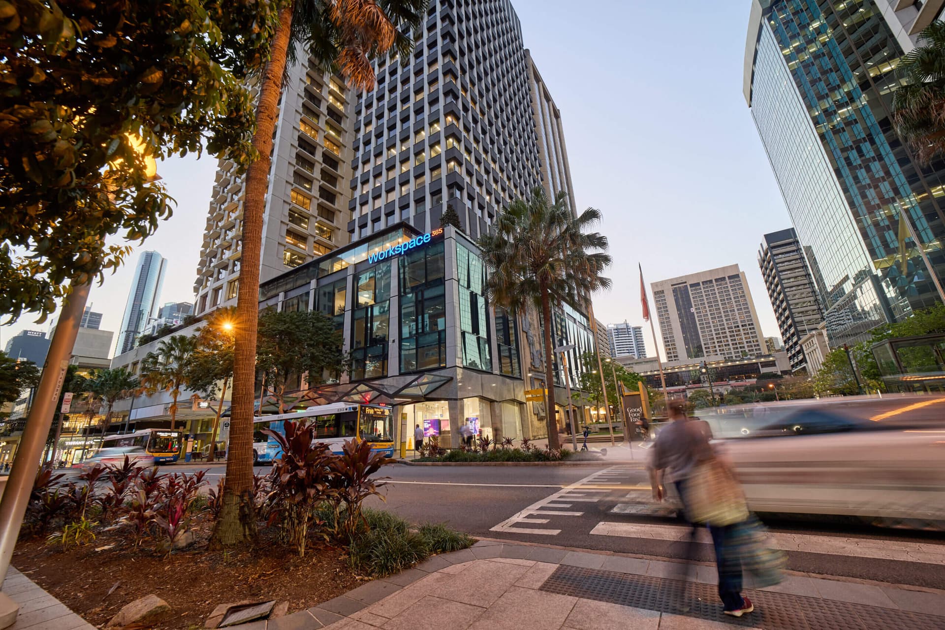 Street scene in Brisbane with a bus, pedestrian crossing, palm trees, and office buildings at dusk.