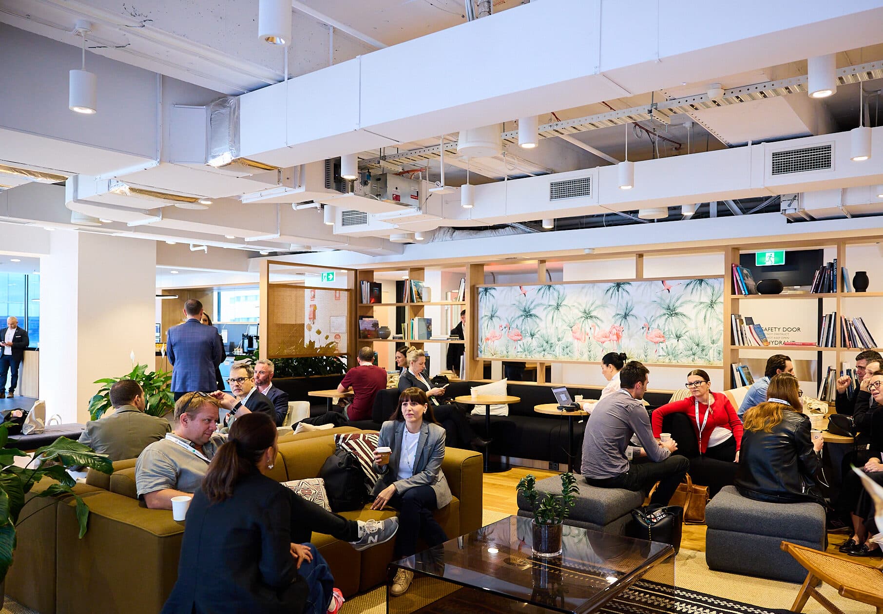 A modern, open-plan office lounge with people sitting on sofas and chairs, engaged in conversations and meetings. Shelves with books, plants, and decorative items line the walls. Bright, welcoming atmosphere.
