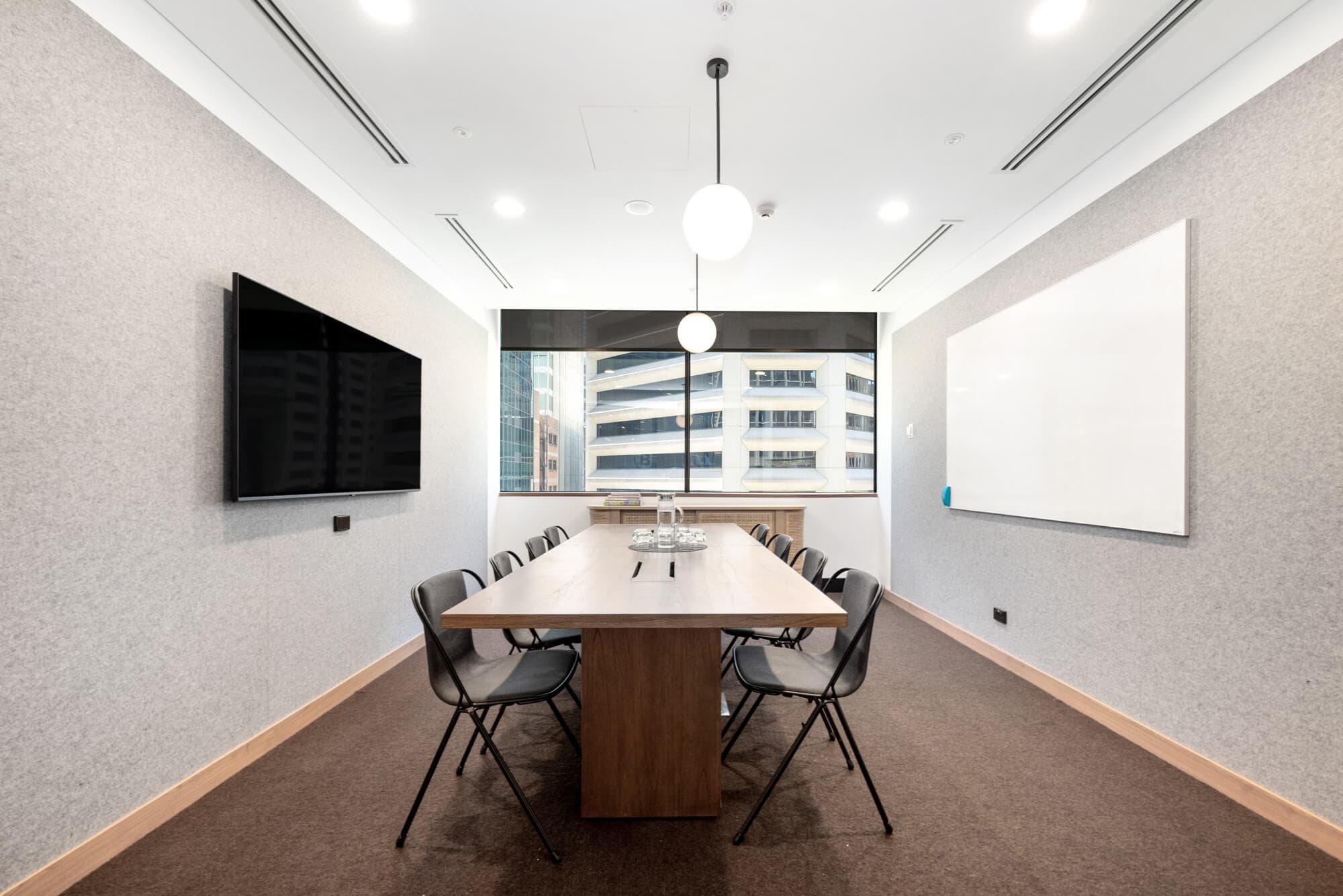 A modern conference room with a long wooden table surrounded by seven black chairs. A large flat-screen TV is mounted on the left wall, and a whiteboard is on the right. The room has overhead lighting and a window with a city view.