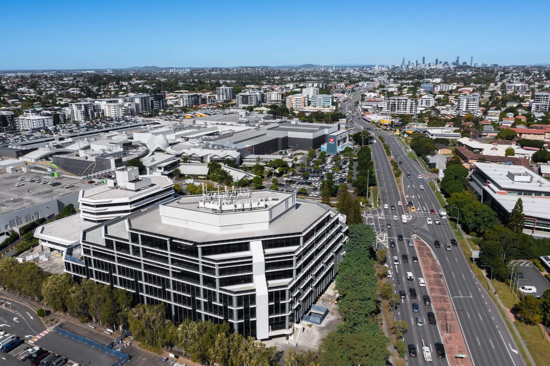 Aerial view of a large urban area with a modern office building in the foreground. Busy streets surround the complex, lined with trees and cars. In the distance, a city skyline is visible under a clear blue sky.