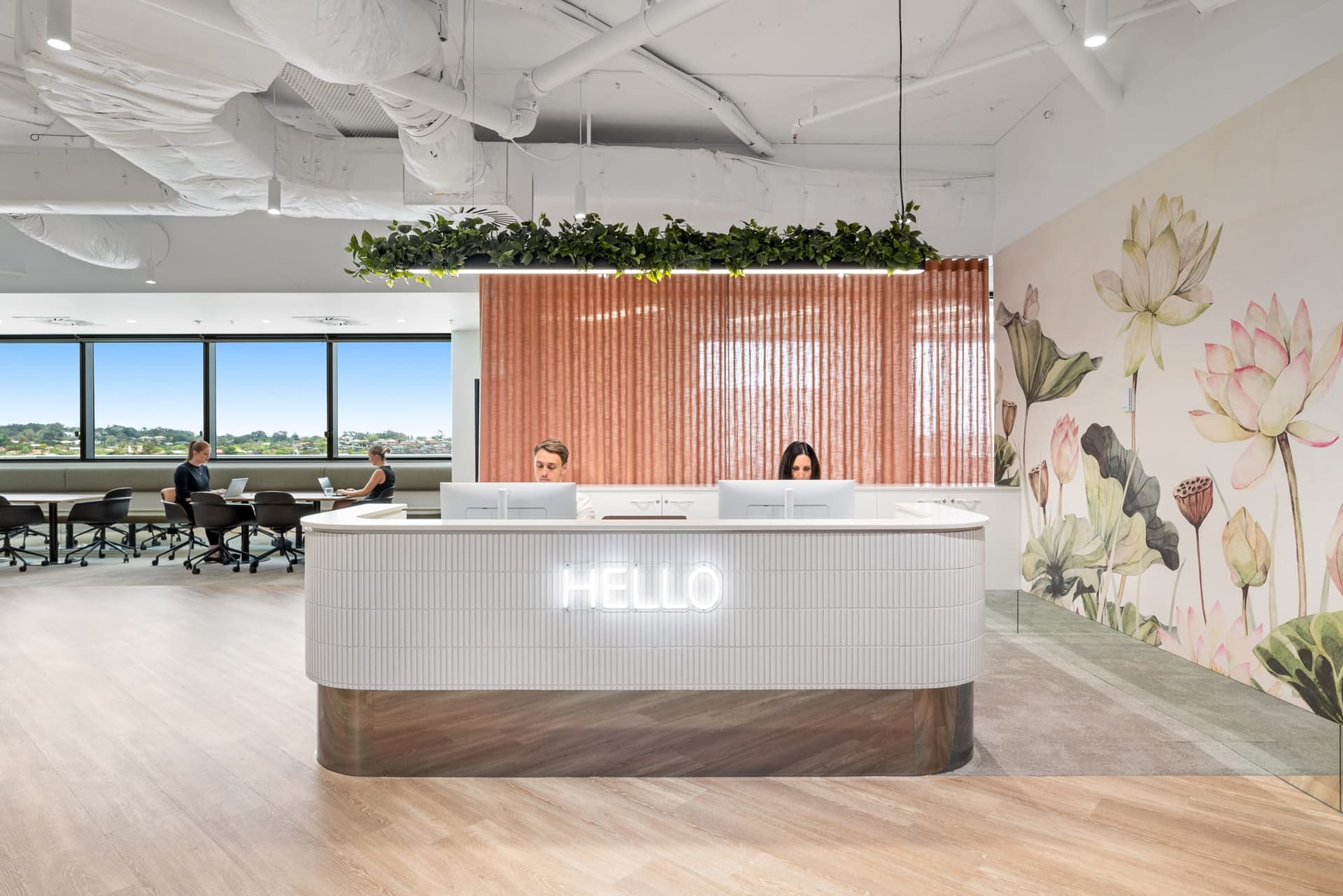 A modern office reception area with two people seated behind a white desk with HELLO illuminated on it. The room features a large window, a lotus-themed mural, and green plants atop the partition. Several people work at tables near the windows.