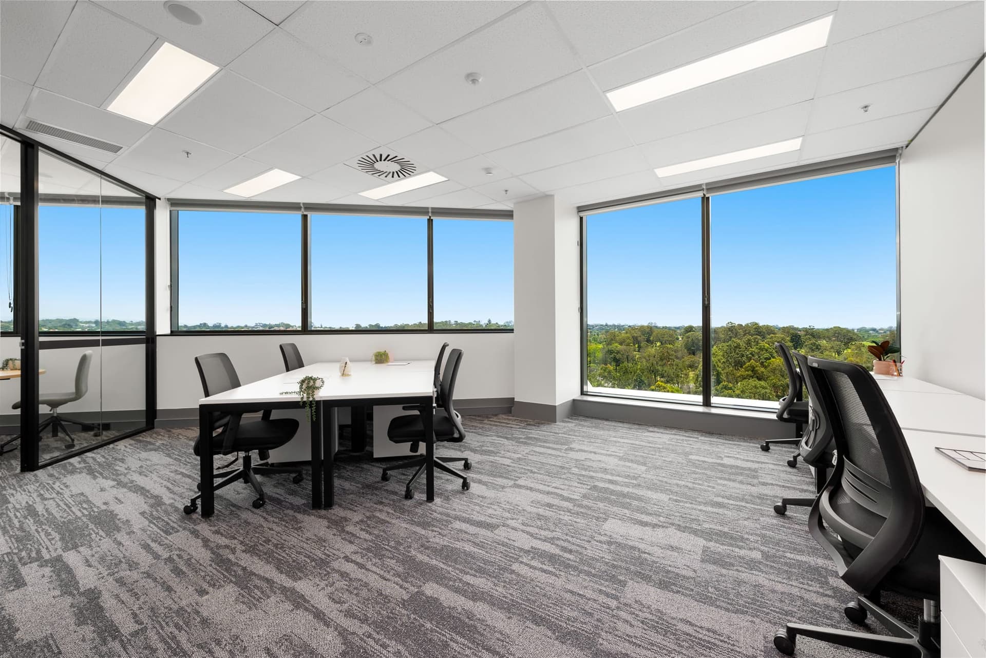 Modern office space with large windows overlooking a green landscape. The room features several desks with black chairs, white walls, and a gray carpet. Natural light fills the room, creating a bright and airy atmosphere.