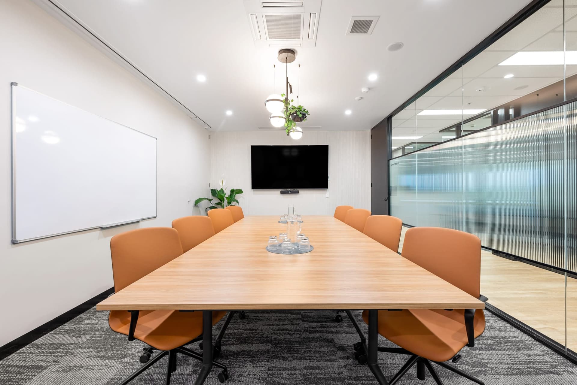 A modern conference room with a long wooden table and eight orange chairs. A large TV is mounted on the front wall, with a whiteboard beside it. The room has glass walls on one side and a potted plant in the corner.