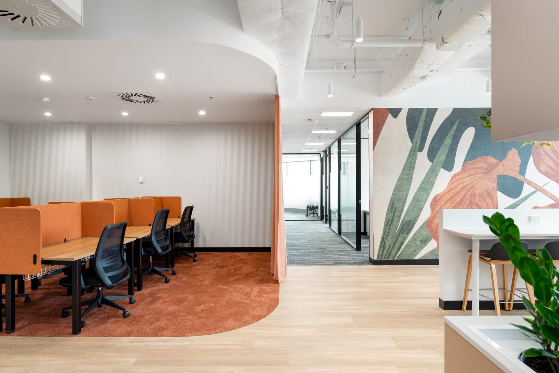 Modern office space with orange cubicle dividers, ergonomic chairs, and a wall mural featuring large leaves. The room has a mix of carpet and wood flooring, with a small plant visible on the right. Ceiling ductwork is exposed.
