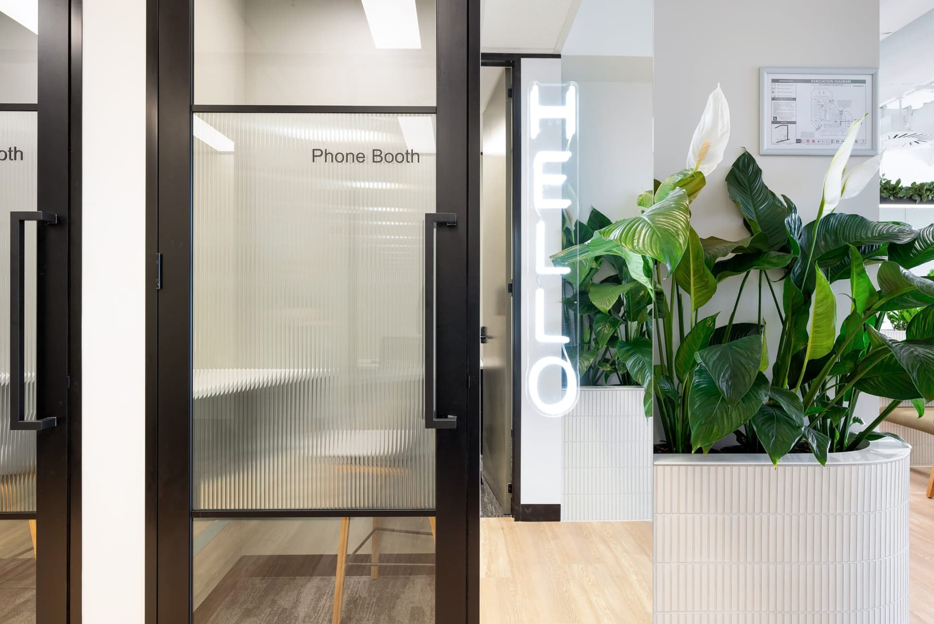 A modern office space with a glass-walled phone booth labeled Phone Booth on the left. To the right is a neon HELLO sign and lush green plants in a white planter. The floor is a mix of wood and carpet.