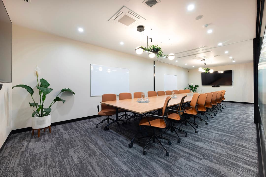 Conference room with large table, brown chairs, whiteboards, TV screen, and indoor plants. Modern, bright office space