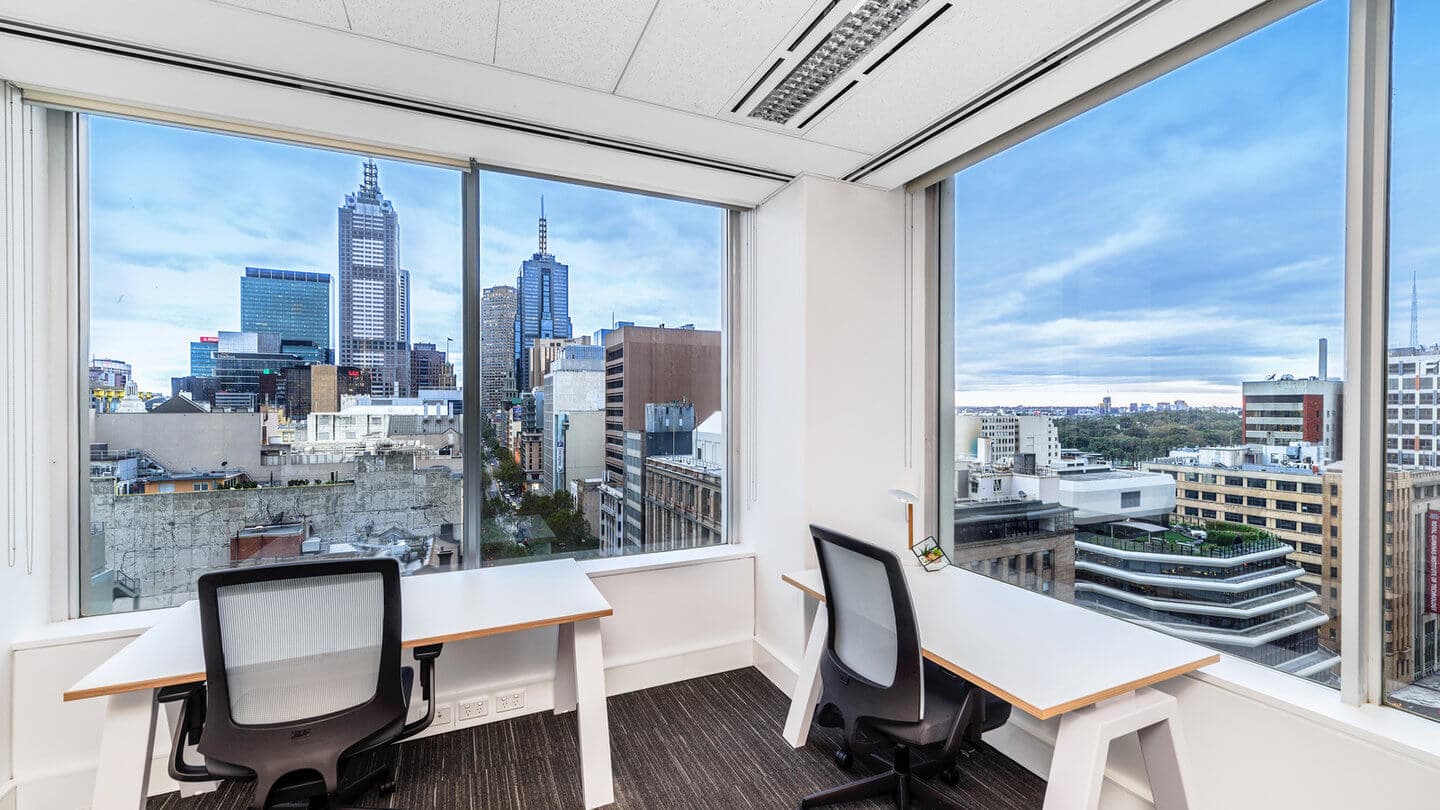 Two empty office desks with chairs facing large windows revealing a cityscape. Skyscrapers and a partly cloudy sky are visible outside. The room has white walls and ceiling, and dark carpet flooring.