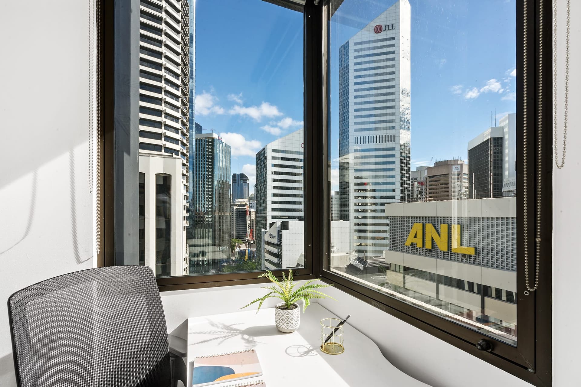 A bright office corner with a desk and chair overlooks a cityscape of tall buildings against a clear blue sky. A small potted plant and a notebook rest on the desk near a window. Prominent building logos visible include ANL and JLL.