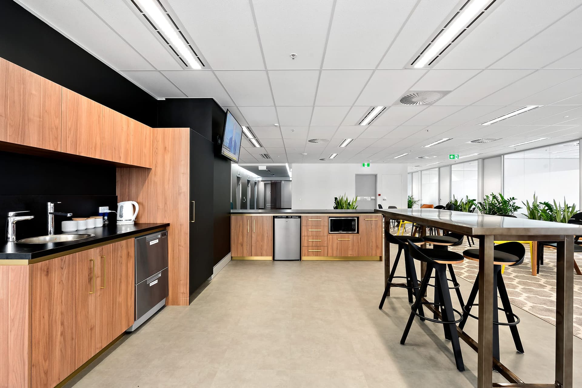 Modern office kitchen with wooden cabinets, stainless steel sink, and appliances including a microwave and kettle. A high counter with black stools sits in the foreground, while potted plants adorn the space. Ceiling lights and large windows brighten the area.