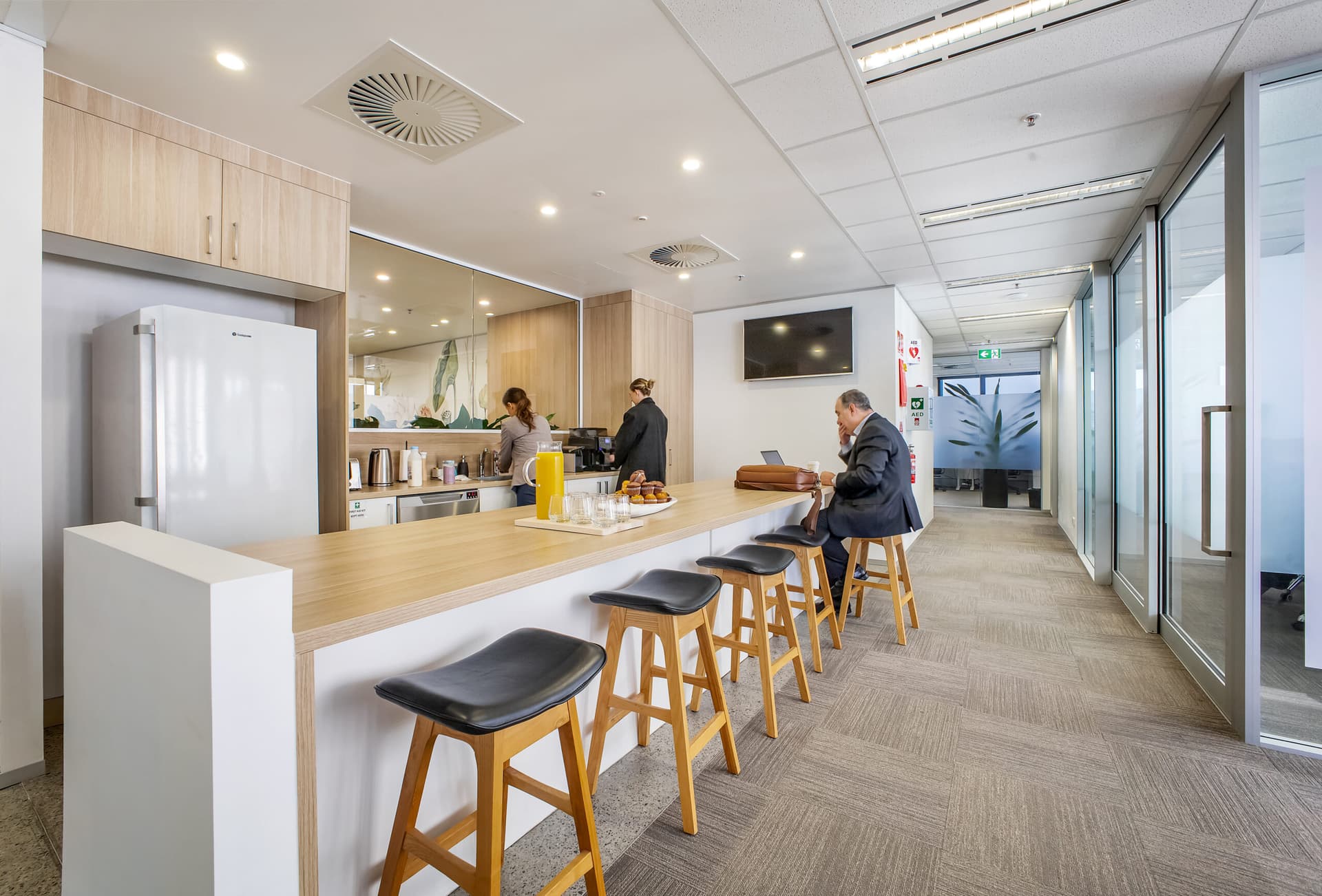Modern office kitchen with a long counter, four bar stools, drinks and pastries on the counter. Two people prepare food in the kitchen area, while one person sits at the counter working on a laptop. Bright and clean environment.