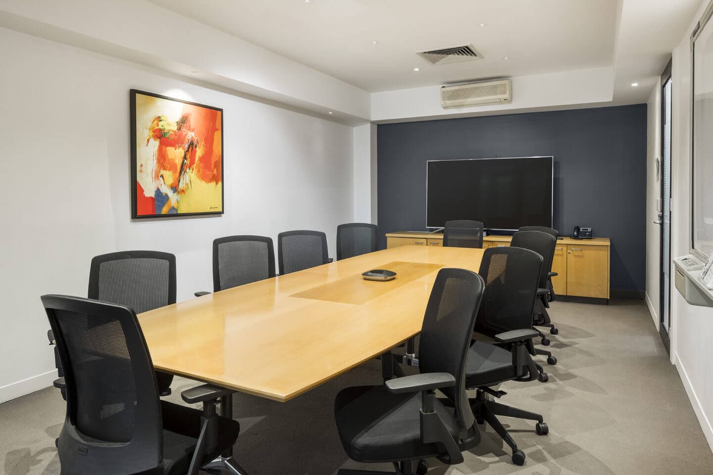A modern conference room with a long wooden table surrounded by eight black mesh office chairs. On the wall, theres an abstract painting with red and yellow tones. A large flat-screen TV is mounted on a dark accent wall. Neutral carpeting covers the floor.