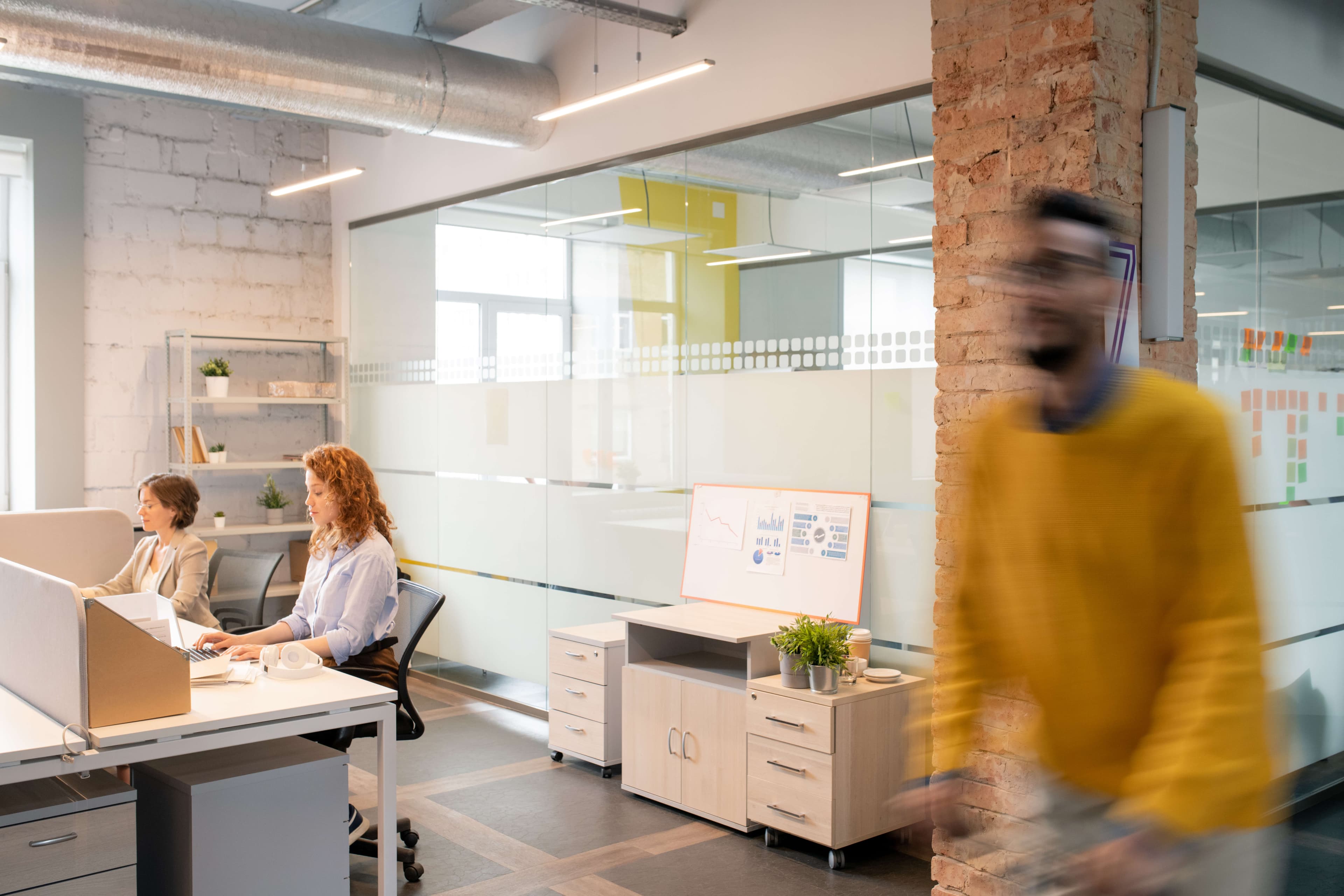 People working in modern open-space office: young ladies sitting at table and using laptops in background, blurred motion of man crossing office