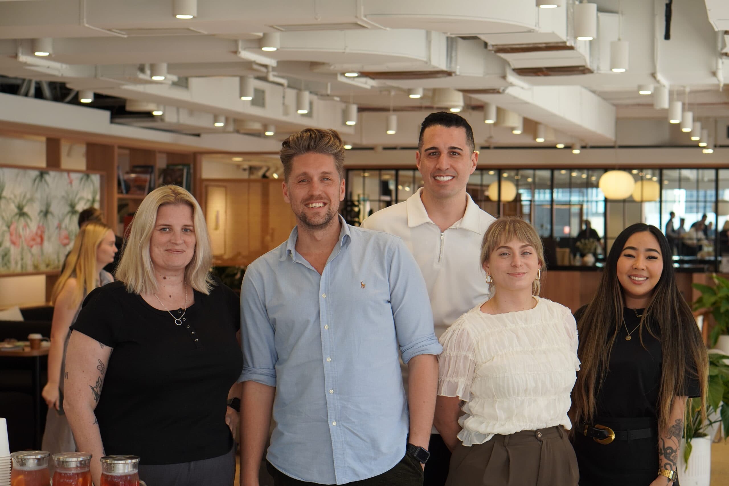 Five people, three women and two men, are standing and smiling together in a modern, well-lit office space share in Sydney, with plants and wooden decor enhancing the vibrant atmosphere.