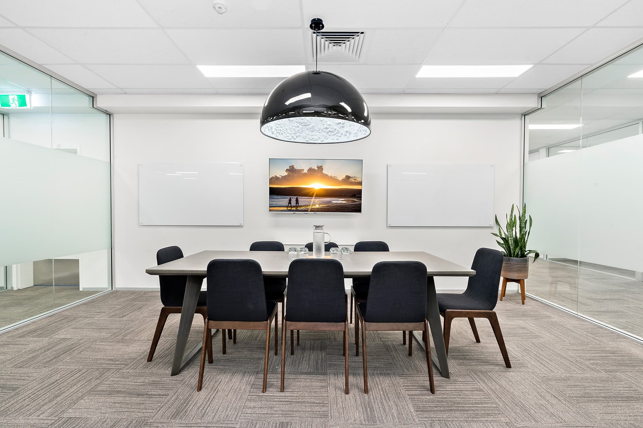 A modern conference room with a large table surrounded by eight dark chairs. A dome-shaped pendant light hangs above. Two blank whiteboards and a landscape photo adorn the walls. A potted plant is in the corner. Glass walls are on both sides.