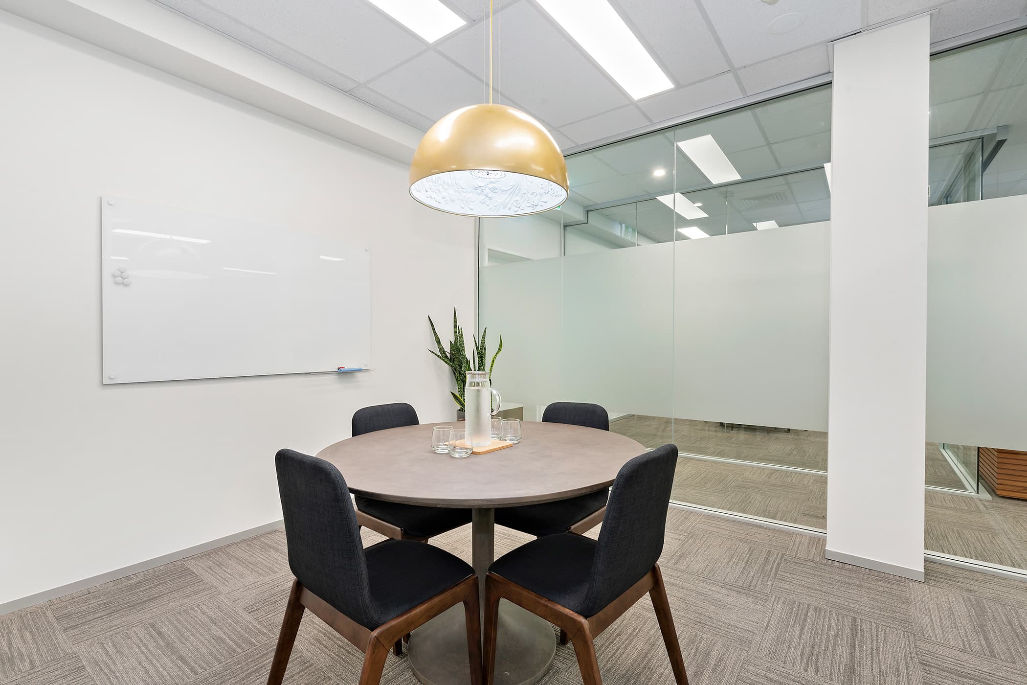 A small conference room with a round table and four black chairs. A large whiteboard hangs on the wall. A gold pendant lamp is above the table, and a potted plant sits in the corner. The room has glass partitions and neutral carpeting.