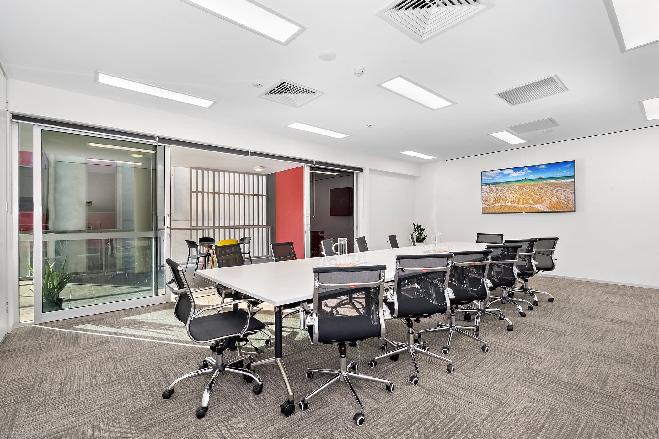 A modern conference room with a long white table surrounded by black office chairs. A large screen displaying a beach scene is mounted on the white wall. Glass doors on the side, allowing natural light to enter.