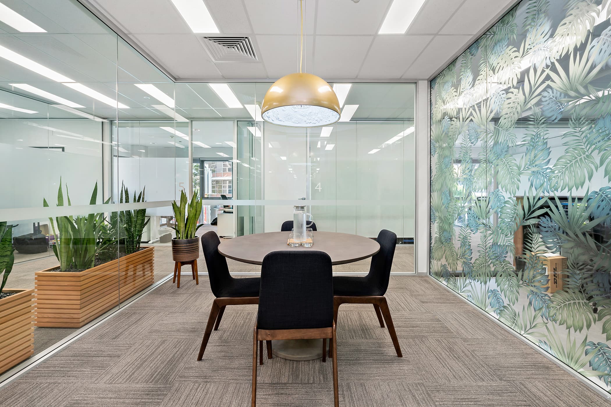 A modern office meeting room with glass walls. A round table is surrounded by four black chairs. A large, gold pendant light hangs above the table. Potted plants are placed in the corner, and one glass wall has a leaf pattern design.