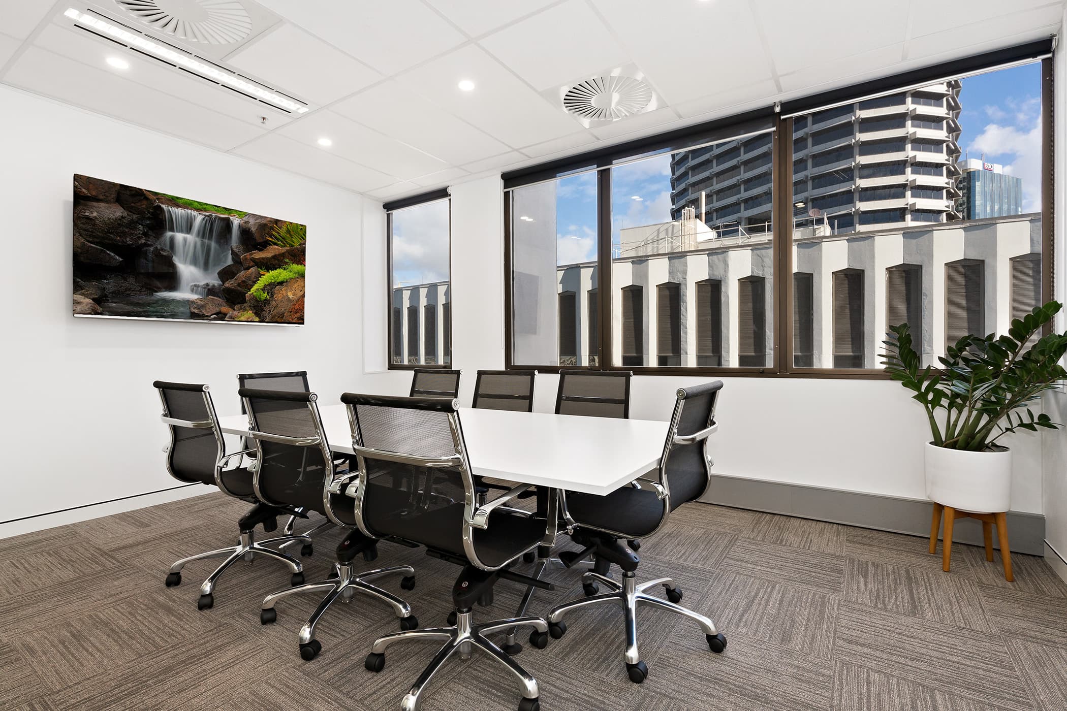 A modern conference room with a long white table surrounded by eight black mesh chairs. A large window displays a city view. A wall-mounted TV shows a waterfall scene. A potted plant is in the corner, and the carpet is gray.