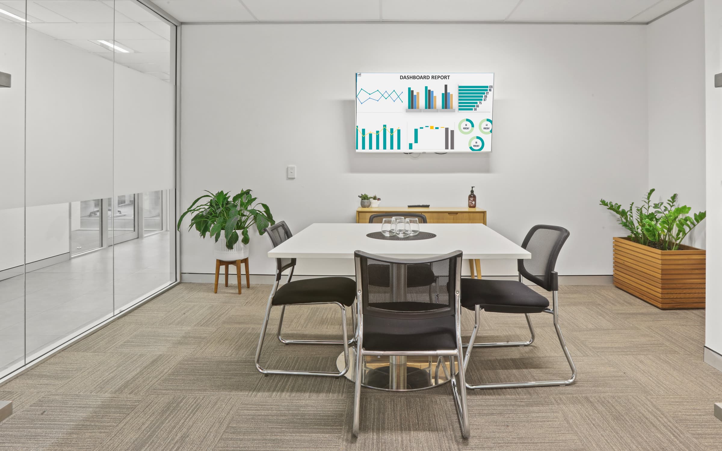 A modern conference room with a white rectangular table, four black chairs, and a digital screen displaying charts. There are two potted plants on the floor. A glass wall on the left separates this room from another space.