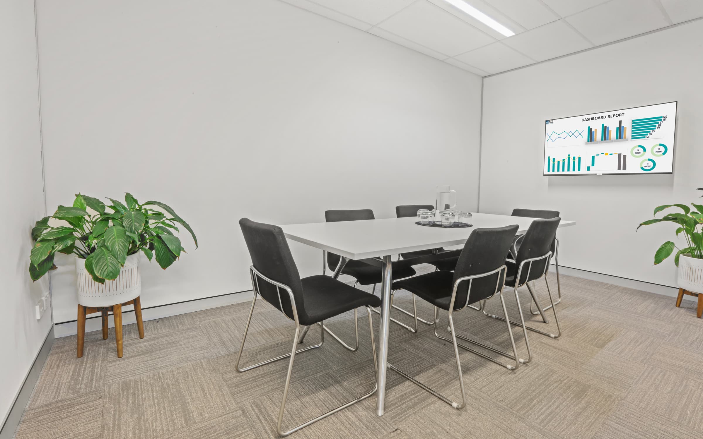 A minimalist conference room with a white table, six black chairs, two potted plants, and a flat-screen displaying graphs on the wall. The room has light gray carpet and white walls, creating a clean and professional environment.