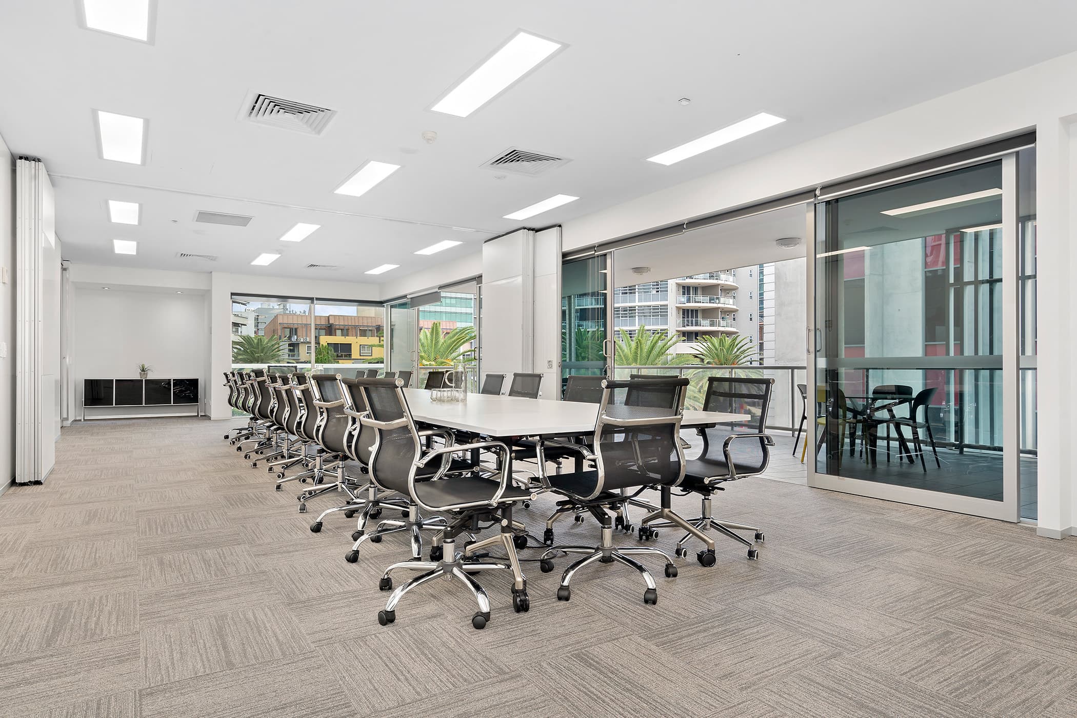 Modern conference room with a long white rectangular table surrounded by black office chairs. Large windows to the right offer cityscape views. The space is well-lit with recessed ceiling lights and features neutral carpet flooring.